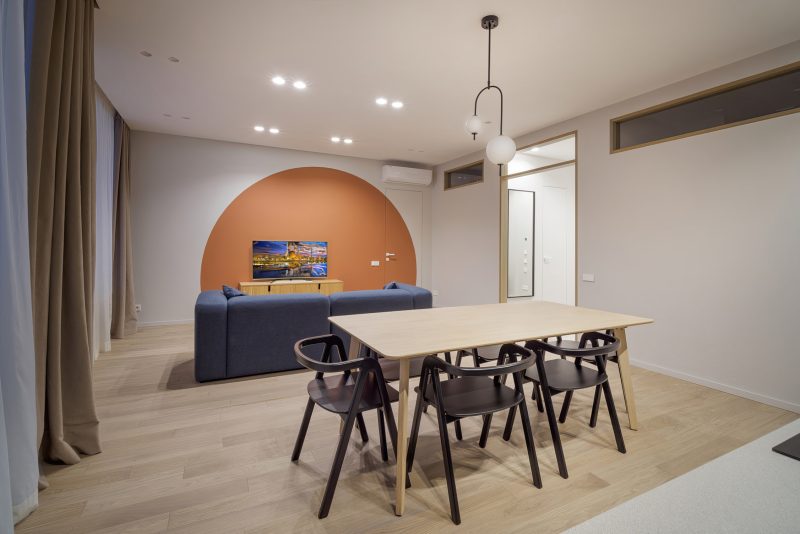 Фотограф Кирилл Толль создает образ интерьера с театральной выразительностью. Полукруг оранжевого цвета напоминает рампу, освещающую сцену с синим диваном. Шарообразные плафоны люстры парят над столом как сферические объекты, их форма повторяется в изгибах стульев. Окно над дверью добавляет воздушности, размывая границы помещения. Светлый паркет отражает общую цветовую гамму, усиливая ощущение простора. Каждый элемент существует в сложном равновесии — массивность стола компенсируется легкостью стульев, яркость акцентной стены уравновешивается спокойствием серого фона. Съемка Кирилл Толль фиксирует момент, когда минимализм обретает эмоциональную глубину.