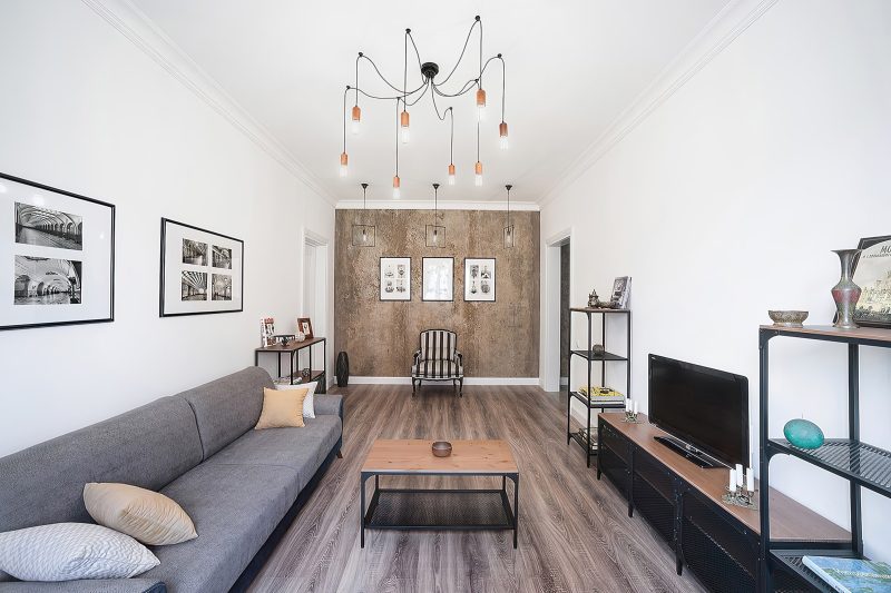 На снимке Кирилла Толль пространство становится философским трактатом. Это не просто гостиная, а поле напряжения между индустриальным прошлым и обжитым настоящим. Фотограф ловит этот момент перемирия: фактурная стена, подобная геологическому пласту, противопоставлена теплой, почти тактильной текстуре деревянного пола. Интерьерная съемка здесь фиксирует не предметы, а материальные аргументы дизайна. Свисающие лампочки — это пунктиры, связывающие высокий потолок с человеческим масштабом низкого столика. Каждый кадр Толль, специалиста по съемке квартир, — это исследование: как грубость, лишенная агрессии, рождает совершенный покой. Мягкая полоска кресла, строгий ряд картин — это знаки оседлости, осторожно вписанные в индустриальный каркас. Пространство дышит историей материалов, где следы времени на стене ценнее любого декора.