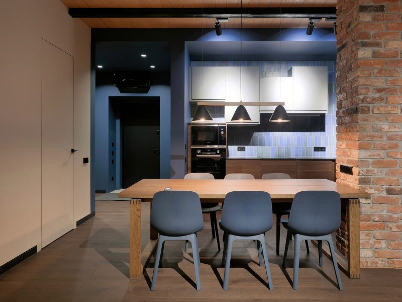 м. Пушкинская. Интерьерная съемка кухни-лофт от фотографа Кирилл Толль: Диалог фактур в Москве. Работа мастера запечатлела пространство, где грубая поэтика кирпичной кладки встречает лаконичный блеск современной кухни. Деревянный стол, массивный, словно вырубленный из цельного ствола, становится эпицентром этого противостояния. Синие стулья вносят дерзкий цветовой акцент, вспышку средиземноморского неба в индустриальном пейзаже. Черные трековые светильники на потолке из досок рисуют жесткую геометрию, подчиняя себе хаос кирпича.