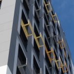 Фрагмент фасада корпуса 3 в ЖК «Новый Зеленоград» (Московская область, Химки, д. Рузино, мкр. Кутузовский, д. 4, корп. 3) . Этот кадр интересен тем, что показывает современную архитектуру не как целостный образ, а как текстурный узор, где каждый элемент работает на общую ритмику. В центре внимания — прямоугольные окна тёмно-серого цвета, расположенные с одинаковым шагом по всей поверхности фасада. Под каждым окном установлен кондиционер — идентичный по форме и размеру, но отличающийся по цвету решётки: серой, голубой или ярко-жёлтой. Этот приём превращает утилитарные элементы в декоративные акценты, разбивая монотонность оконного ряда и добавляя индивидуальность каждому жилому блоку. Вертикальные полосы, разделяющие секции фасада, выполнены в белом, светло-сером, тёмно-сером и бежево-жёлтом цветах. Слева фрагмент фасада имеет более монохромную палитру, справа — яркие жёлтые и оранжевые элементы создают визуальное оживление. Здание облицовано фиброцементными плитами — экологичным материалом, который обеспечивает долговечность и современный внешний вид . Съёмка велась в дневное время при рассеянном свете, без резких теней. Это позволило равномерно осветить фасад и точно передать все цветовые нюансы. Отсутствие неба и земли в кадре фокусирует внимание исключительно на архитектурной геометрии и деталях. Фотография демонстрирует, как продуманное цветовое решение и внимание к мелочам формируют уникальный облик современного жилого комплекса, спроектированного бюро SPEECH