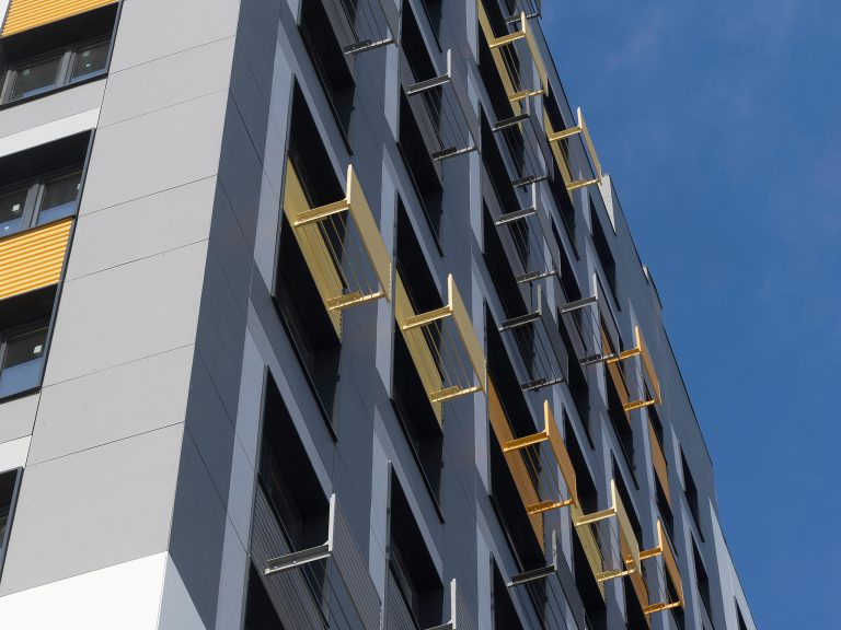 Фрагмент фасада корпуса 3 в ЖК «Новый Зеленоград» (Московская область, Химки, д. Рузино, мкр. Кутузовский, д. 4, корп. 3) . Этот кадр интересен тем, что показывает современную архитектуру не как целостный образ, а как текстурный узор, где каждый элемент работает на общую ритмику. В центре внимания — прямоугольные окна тёмно-серого цвета, расположенные с одинаковым шагом по всей поверхности фасада. Под каждым окном установлен кондиционер — идентичный по форме и размеру, но отличающийся по цвету решётки: серой, голубой или ярко-жёлтой. Этот приём превращает утилитарные элементы в декоративные акценты, разбивая монотонность оконного ряда и добавляя индивидуальность каждому жилому блоку. Вертикальные полосы, разделяющие секции фасада, выполнены в белом, светло-сером, тёмно-сером и бежево-жёлтом цветах. Слева фрагмент фасада имеет более монохромную палитру, справа — яркие жёлтые и оранжевые элементы создают визуальное оживление. Здание облицовано фиброцементными плитами — экологичным материалом, который обеспечивает долговечность и современный внешний вид . Съёмка велась в дневное время при рассеянном свете, без резких теней. Это позволило равномерно осветить фасад и точно передать все цветовые нюансы. Отсутствие неба и земли в кадре фокусирует внимание исключительно на архитектурной геометрии и деталях. Фотография демонстрирует, как продуманное цветовое решение и внимание к мелочам формируют уникальный облик современного жилого комплекса, спроектированного бюро SPEECH