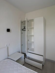 Белый трехстворчатый шкаф с открытыми дверцами, выдвинутыми ящиками и пустой штангой в спальне.