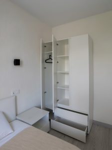 Открытый белый шкаф рядом с кроватью, застеленной сгенерированным бежевым покрывалом и подушкой.