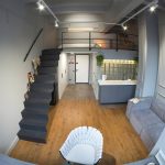 Вид сверху на двухуровневые апартаменты в ЖК Loftec: антресольный этаж со спальней, чёрная лестница, серая кухня с фартуком из стеклоблоков и трековое освещение — Кирилл Толль