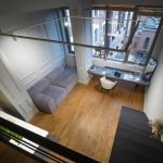 Вид сверху на гостиную зону в ЖК Loftec с серым модульным диваном, встроенным рабочим столом у панорамного окна и чёрной металлической лестницей — фото Кирилла Толль