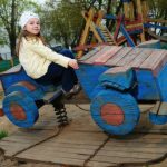 Фотография запечатлела маленькую девочку, сидящую на самодельной деревянной игрушке в форме автомобиля на игровой площадке, в окружении других детских игровых конструкций.

Композиция кадра построена таким образом, что главная героиня, девочка, находится в центре внимания, занимая правую половину изображения. Она сидит на ярко-синем деревянном игрушечном автомобиле, который занимает большую часть переднего плана. Автомобиль установлен на деревянной платформе, от которой отходят цепи, ведущие к пружинному механизму, что указывает на его качающуюся конструкцию. Взгляд девочки направлен прямо в камеру, что создает ощущение прямого контакта со зрителем. На заднем плане видны элементы более крупной детской площадки, выполненной из дерева, а также деревья и, вдалеке, мужчина, сидящий на скамейке, что дополняет атмосферу общественного пространства.

Ключевой субъект – девочка, возрастом примерно 7-8 лет, с длинными светлыми волосами, заплетенными в две косички. На голове у нее белая повязка с рисунком. Девочка одета в желтую ветровку, темно-синие штаны и коричневые детские кроссовки. Выражение ее лица спокойное, с легкой улыбкой, что передает ощущение уверенности и комфорта. Она сидит, держась одной рукой за борт игрушечного автомобиля. Игрушечный автомобиль, являющийся вторым важным элементом, сделан из грубо обработанных деревянных досок, окрашенных в ярко-синий цвет. Колеса автомобиля также деревянные, с красными акцентами. Конструкция выглядит самодельной, что придает ей особый шарм и аутентичность.

Этот снимок выполнен в жанре уличной фотографии, с акцентом на естественность момента. Техническое исполнение фотографии находится на высоком уровне: четкое изображение, правильная экспозиция и сбалансированный баланс белого. Стиль фотографии реалистичный, без излишней ретуши или художественных фильтров. Цветовая гамма снимка состоит из природных оттенков, с преобладанием синего, желтого и землистых тонов, что характерно для изображения уличной детской площадки. Креативный выбор заключается в фиксации повседневного момента из жизни ребенка, подчеркивая его радость и увлеченность игрой.

Сцена разворачивается на открытой игровой площадке, с песчаным покрытием, что типично для многих детских зон. Освещение в кадре естественное, дневное, рассеянное, что свидетельствует о пасмурном дне или съемке в тени деревьев. Это освещение мягко ложится на объекты, не создавая резких теней и подчеркивая текстуру дерева. На заднем плане видны зеленые кроны деревьев, что указывает на весенний или летний период. Общая атмосфера снимка – спокойствие, умиротворение и детская беззаботность. Окружающие элементы, такие как другие части игровой площадки и сидящий вдалеке мужчина, создают контекст для основной сцены, делая ее частью более широкого городского пейзажа.