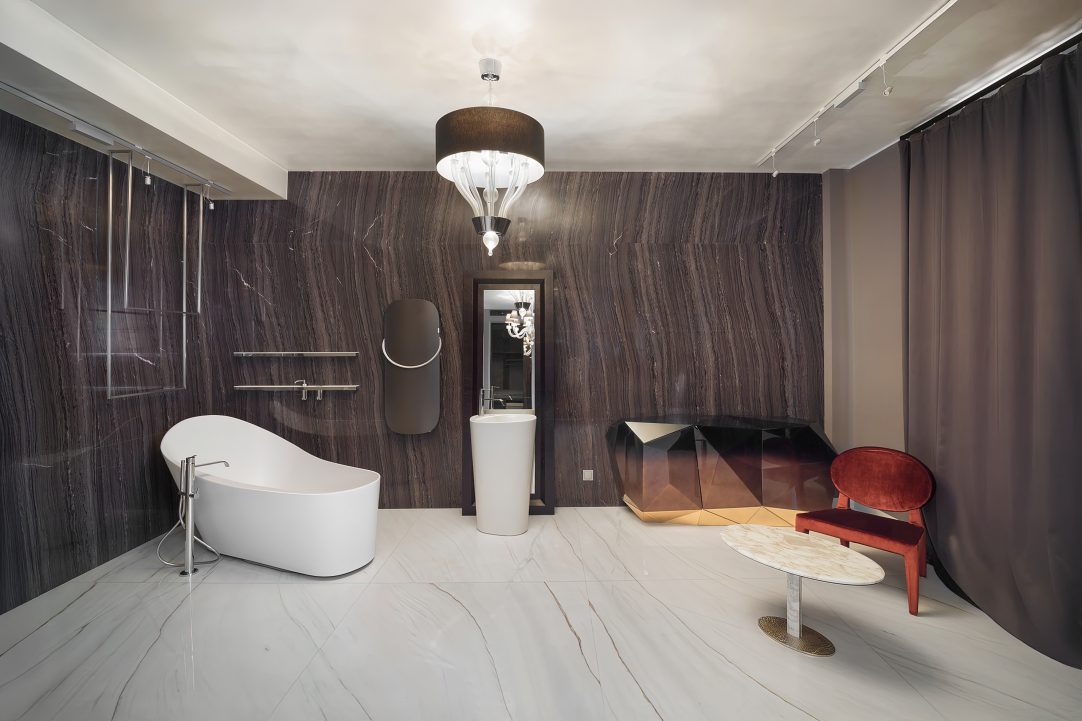 Роскошная ванная комната с отделкой стен темным натуральным камнем, отдельно стоящей дизайнерской ванной, зеркальным комодом и красным бархатным креслом.