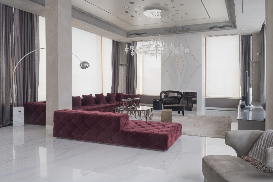 Просторная гостиная в стиле ар-деко с панорамными окнами, бордовым модульным диваном Longhi Must, хрустальной каскадной люстрой и белым мраморным полом.