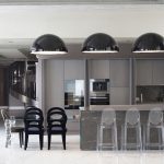 Современный интерьер кухни-гостиной в пентхаусе с кухонным островом из темного мрамора, черными дизайнерскими стульями Promemoria Bilou Bilou и прозрачными барными стульями Kartell под тремя черными купольными светильниками.