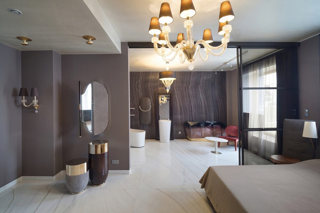 Интерьер мастер-спальни с открытой ванной комнатой, разделенных стеклянной перегородкой, с люстрой в классическом стиле, отдельно стоящей сантехникой и стеной из натурального темного камня.