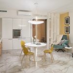 Интерьер кухни-гостиной открытой планировки в ЖК Сады Пекина с белым гарнитуром, круглым столом, жёлтыми стульями, бирюзовым креслом и мраморным полом — съёмка Кирилла Толль
