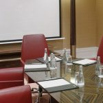 Вертикальный ракурс стола из полосатого шпора зебрано, кожаных папок-бюваров, эргономичных красных кресел, низкой тумбы темного дерева, высокого цилиндрического торшера и рулонного экрана проектора
