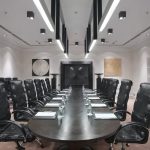 Строгий продольный кадр стола заседаний Radisson Royal с разложенными кожаными бюварами, черными креслами, квадратной рельефной картиной золотистого оттенка и встроенным в глубокую нишу телевизором