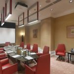 Широкий ракурс конференц-зала Radisson Royal с прямоугольным глянцевым столом зебрано, красными кожаными стульями, белым проектором Panasonic под потолком и золотисто-охристыми текстурными обоями