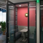 Изолированная коралловая переговорная капсула VK Meeting Pod с открытой стеклянной дверью, фетровой отделкой стенок, зеленым бра и ковролином.