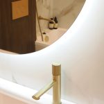 Деталь ванной комнаты в ЖК Level Причальный: матовый золотой смеситель, круглое зеркало с мягкой подсветкой.