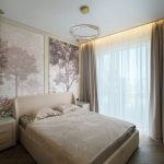 Интерьер светлой спальни в ЖК Level Причальный: кровать с бежевой обивкой, настенное панно с лесом, кольцевая люстра с хрусталем.