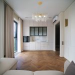 Фронтальный вид кухни-гостиной в ЖК Level Причальный: белый кухонный гарнитур, паркет елочка, дизайнерская хрустальная люстра.