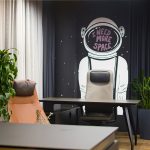 Рабочее место с эргономичным компьютерным креслом графитовым столом черной матовой стеной с белой графикой космонавта серым кашпо замиокулькас плотными портьерами