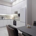Угловая белая кухня с глянцевыми фасадами в ЖК Мосфильмовский. Фартук под мрамор, встроенная подсветка, белый холодильник и обеденный стол. Мосфильмовская 88к4с2.