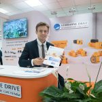 Работа стендиста на промышленной выставке, панорамный кадр выставочной зоны с моделью в деловом костюме и оранжевыми редукторами