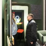 Вертикальный репортажный кадр высокой концентрации. Профили сотрудницы с выставочным бейджем и клиента в момент серьезного обсуждения проекта у открытой стеклянной двери кабины. Внутренний неоновый знак желтого цвета From SPACE with ♡ создает эффектный контровой световой контур.