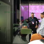 Архитектурно-репортажный кадр. Рустем Мамин ведет дискуссию с архитектором на стыке переговорной группы и массивного корпуса акустической кабины SPACELINE, выступающей левой кулисой кадра. На дальнем плане открывается глубокая и размытая панорама соседних выставочных павильонов.