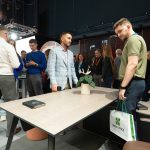 Кадр тактильного контакта с продуктом. Посетитель в оливковой футболке, удерживая в руке фирменный бумажный пакет Space Office с зеленым логотипом, внимательно рассматривает матовую текстуру деревянного шпона на столе руководителя. На столе разложены черные буклеты DETAILS MATTER.