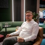 Портретная съемка с малой глубиной резкости. Денис Силавин в очках и светлой рубашке сидит вполоборота в мягком кресле лаунж-зоны, удерживая в руке стакан с кофе. Мгновенная фиксация расслабленной позы. На заднем плане размыты ритмичные вертикальные световые полосы зеленой стены стенда.