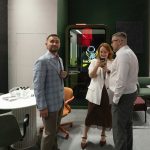 Репортажная фиксация цифрового взаимодействия. Рустем Мамин, Денис Медведев и сотрудница в белом жакете стоят плотной группой у кабины тишины и сосредоточенно изучают информацию на экране смартфона. Баланс теплого интерьерного света и глубокого зеленого цвета стен застройки.