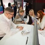 Художественный репортажный прием. Смазанный в движении силуэт спикера на переднем плане слева передает высокую скорость процессов выставки. В резкости находятся улыбающиеся лица гостей и менеджер в белом жакете, демонстрирующая кабель-менеджмент на белой столешнице.