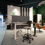 Технический кадр умного рабочего места Sit-to-Stand линейки SPACELINE. Белый металлический каркас стола с электронной системой подъема столешницы, плотный серый звукоизоляционный экран из фетра и высокий регулируемый стул-седло на колесиках с круглой подставкой для ног. Виден гибкий кабель-канал змея.