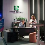 Кинематографичный кадр рабочего взаимодействия. Денис Медведев сидит за столом руководителя и жестикулирует в процессе веселого диалога с сотрудницей. Девушка стоит в профиль с блокнотом в руках. Передний план слева деликатно обрамлен размытым краем серого акустического экрана стола.