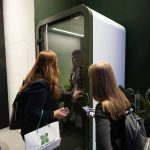 Взгляд через плечо гостей. Две девушки-посетительницы внимательно разглядывают внутреннее устройство кабины тишины SPACELINE. В руках одной из них — фирменный пакет Space Office с ярко-зеленым логотипом. Изнутри кабины через стекло им открыто улыбается менеджер компании.