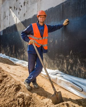 Парень-строитель в каске с лопатой, позирует для рабочего портрета на фоне стройки