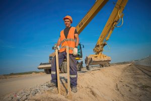 Молодой рабочий в каске стоит на краю дороги, портрет на фоне техники