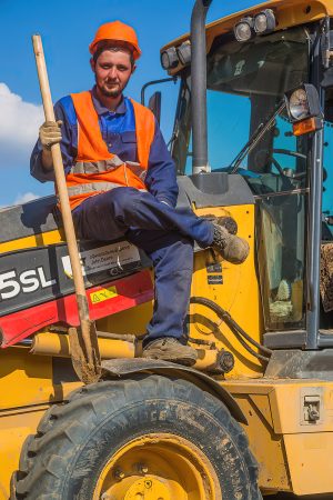 Студент-геодезист в оранжевом жилете отдыхает, сидя на тракторе, живой репортажный портрет